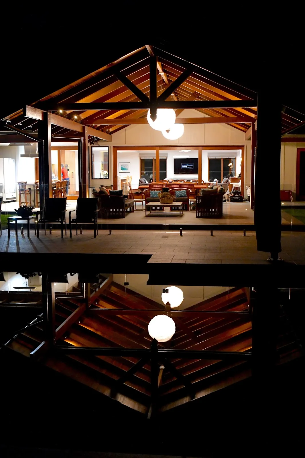 Outdoor timber pavilion at night with downlights and pendant globes reflected in the pool